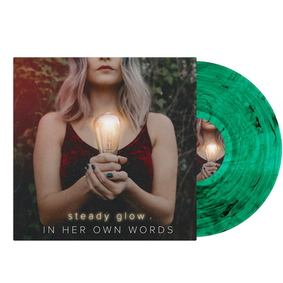 Image of the in her own words steady glow vinyl sleeve with a green and black smoke effect vinyl sticking halfway out of the sleeve. The background is transparent. The album artwork is an image of a woman standing in front of leaves and branches, photographed from her nose down to her midsection. She has short, light pink hair and is wearing a red velvet dress. She holds a glowing lightbulb. Below that in glowing yellow and white font reads "steady glow" "in her own words".